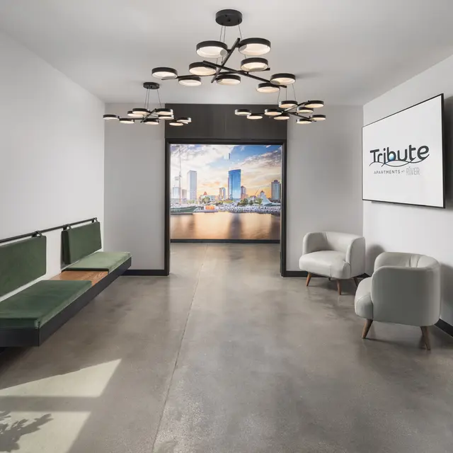 A modern entryway featuring a green bench and contemporary lighting. A digital display is visible at the back with a cityscape image. There are two white chairs and a large plant near the entrance.