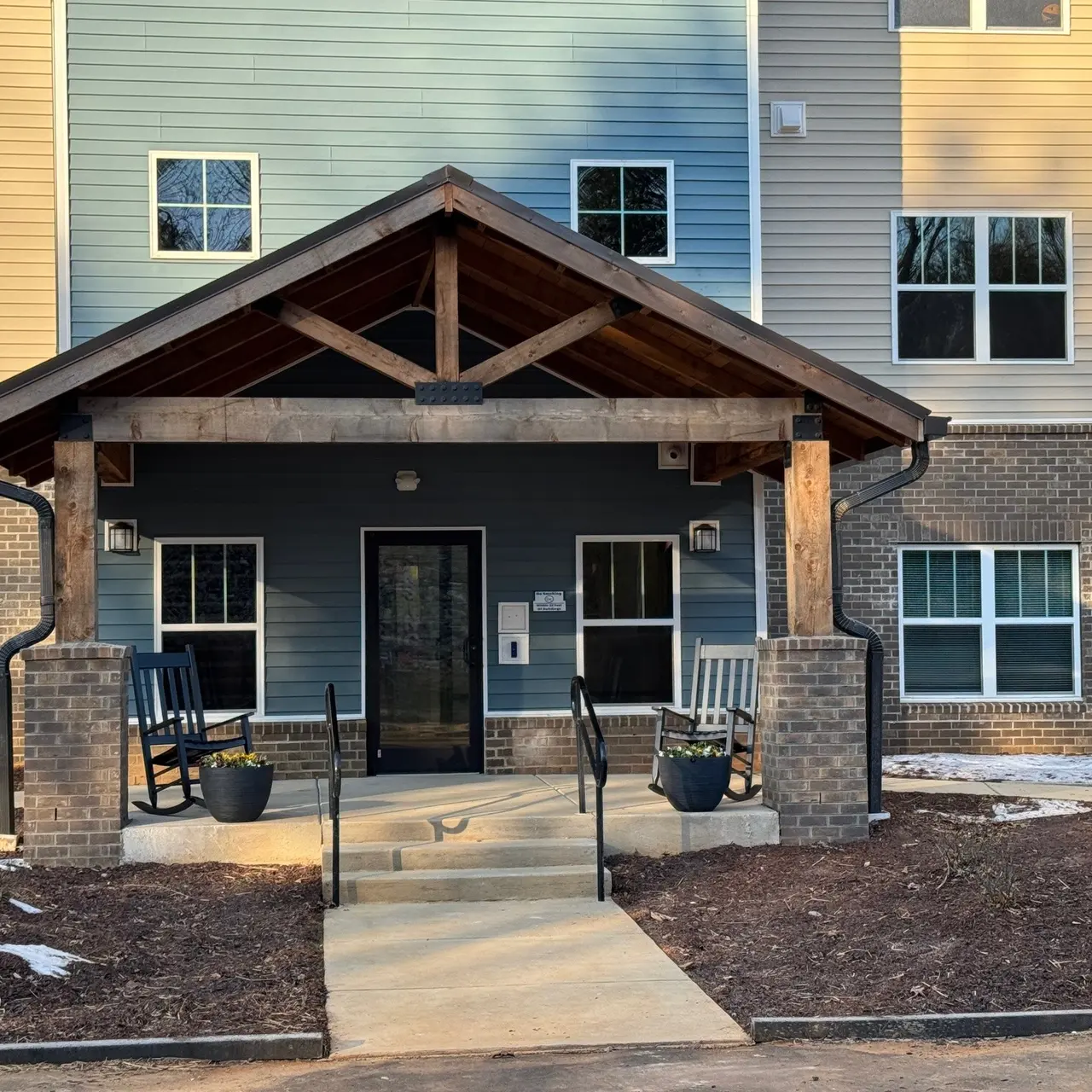 Entrance of an apartment building featuring a covered porch, rocking chairs, and a ramp.