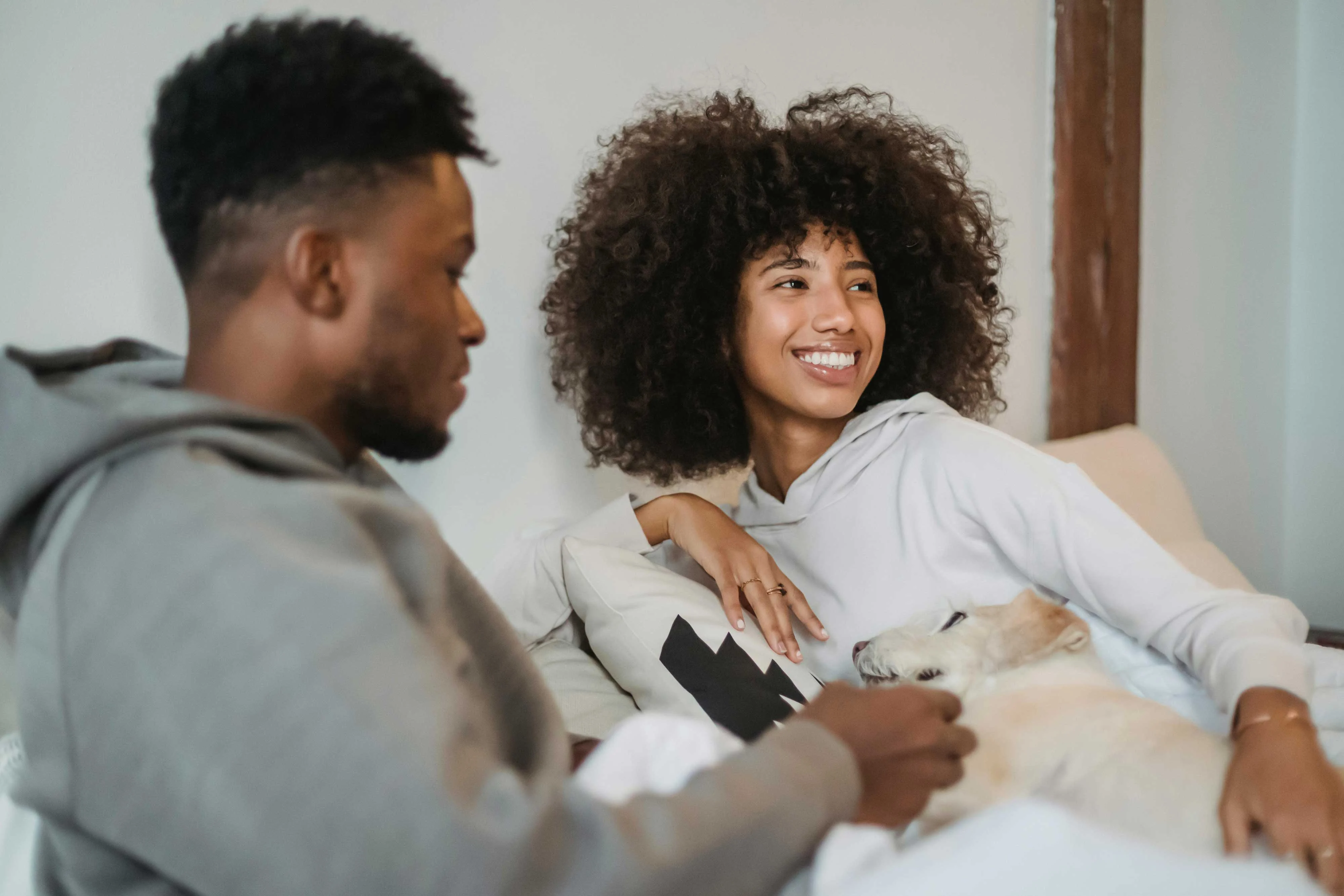 A couple sitting together on a couch, smiling and enjoying each other's company. A small dog is lying comfortably beside them.