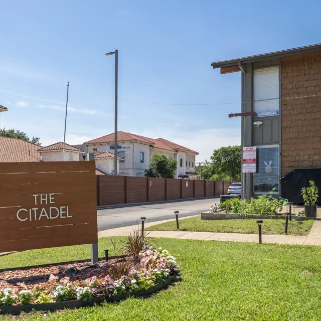 Exterior view of The Citadel apartment complex, featuring a wooden sign and surrounding grassy area with flowers.