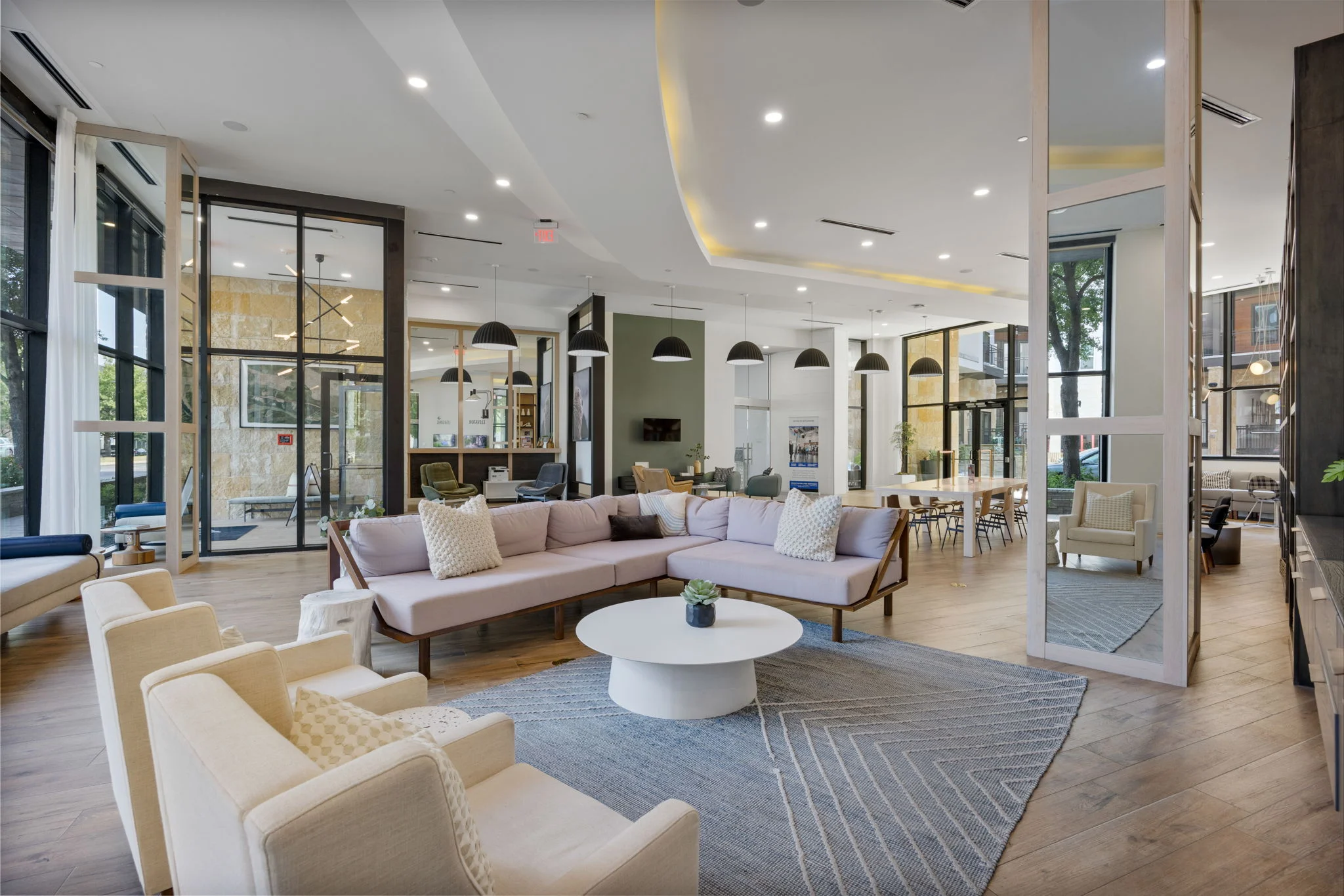 A spacious modern living area with large windows, contemporary furniture, and natural light. There is a sectional sofa with cushions, a round coffee table, and several seating areas. The decor includes a patterned rug and stylish light fixtures.