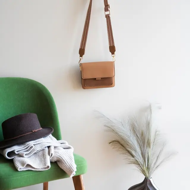 A stylish arrangement of a brown crossbody bag hanging on a wooden hook, with a green chair, a gray sweater folded on the seat, a black fedora hat resting on top, and a decorative leaf-patterned vase holding decorative grasses.