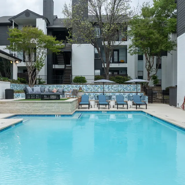 A spacious outdoor swimming pool area featuring clear blue water surrounded by a tan deck. Comfortable lounge chairs are set up near the pool, and lush greenery and trees enhance the ambiance. Apartment buildings are visible in the background.