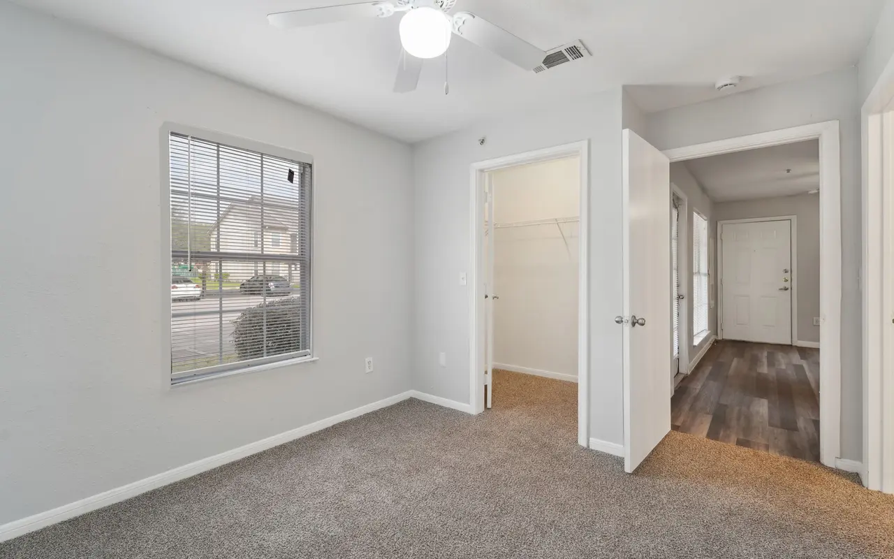 A cozy bedroom featuring carpeted flooring, a ceiling fan, and a window allowing natural light. There is a closet with sliding doors and an adjacent hallway leading to the entrance.