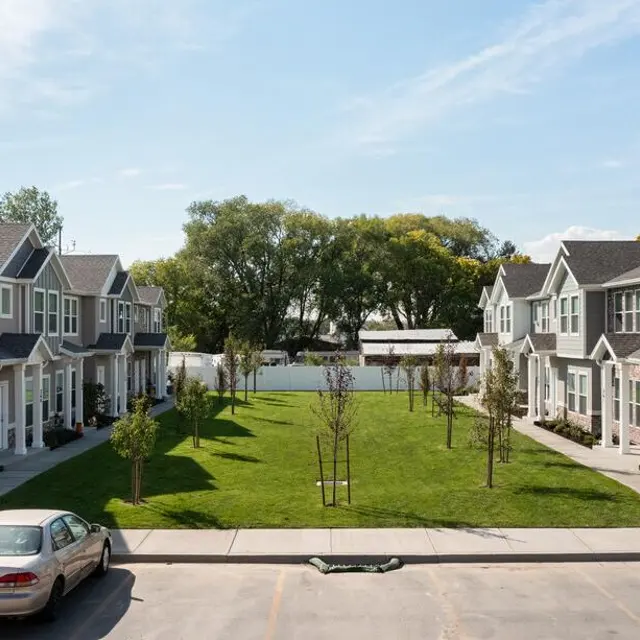 View of a modern apartment complex with green grass in the center and parked cars in front.