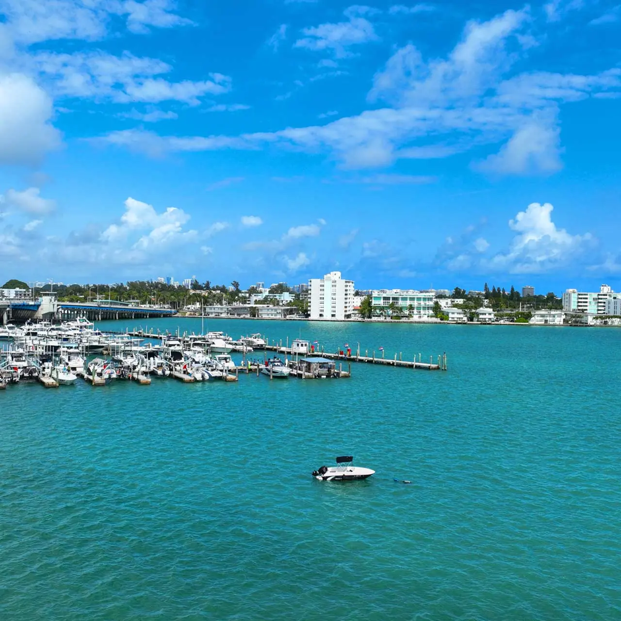A scenic view of a marina with numerous boats docked, surrounded by a calm turquoise sea under a blue sky with fluffy white clouds.