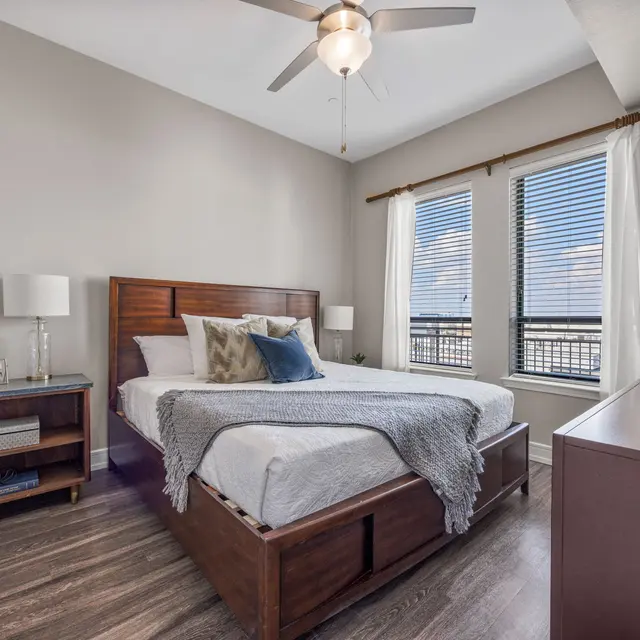 A stylish bedroom featuring a wooden bed frame with a light gray blanket and decorative pillows, two windows with white curtains allowing natural light, a bedside lamp, and a wooden dresser.