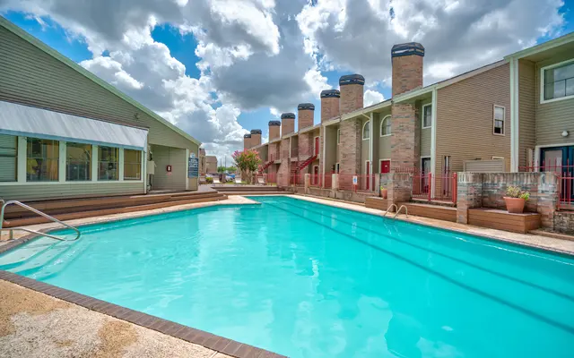 A swimming pool surrounded by apartment buildings with tall chimneys in the background. The pool is clear and inviting, with lounge chairs and patios nearby. The sky is partly cloudy, creating a bright atmosphere.
