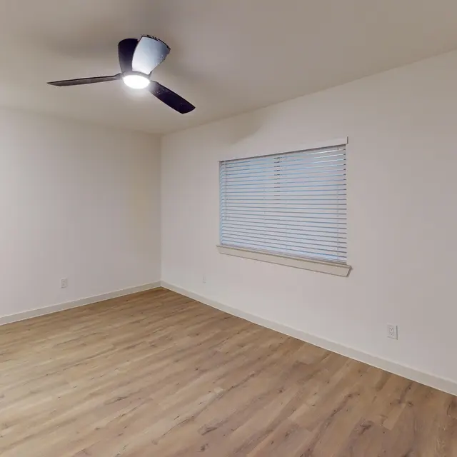 An empty room with light-colored walls and wood-like flooring. A ceiling fan is mounted above, and there is a window with blinds on one wall.