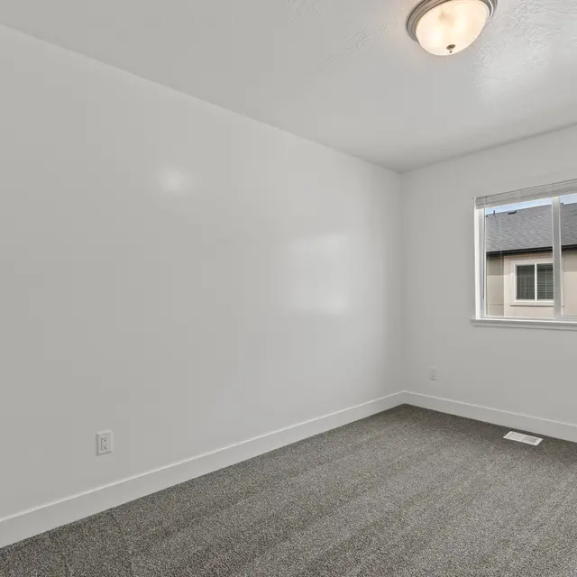 An empty room with grey carpet, white walls, and a window. A light fixture is visible on the ceiling.