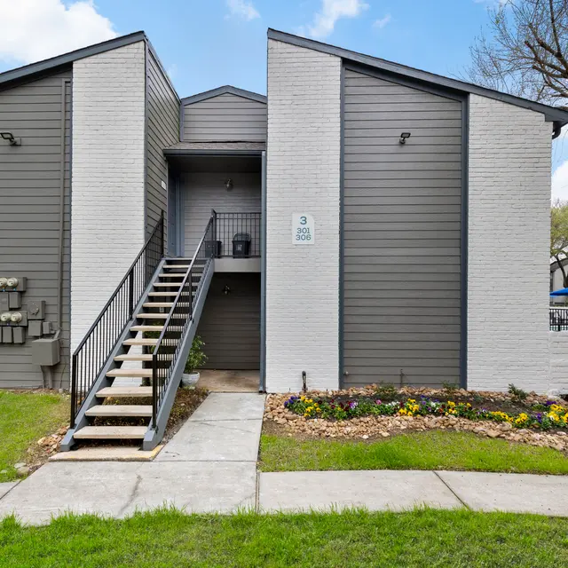 Exterior View of Apartment Building Exterior view of a two-story apartment building with grey and white siding, featuring a staircase leading to the entrance and colorful flower beds.