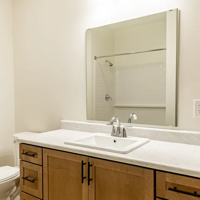 A modern bathroom featuring a clean, minimalist design. It includes a white sink with a stylish faucet on a light-colored countertop, a wooden cabinet with drawers, a mirror above the sink, and a glass shower enclosure in the background. The walls and floor are finished in neutral tones, providing a bright and airy feel.