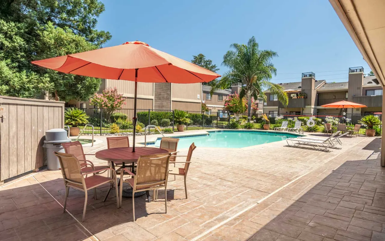 A sunny outdoor swimming pool area featuring a pool, lounge chairs, and tables with umbrellas.
