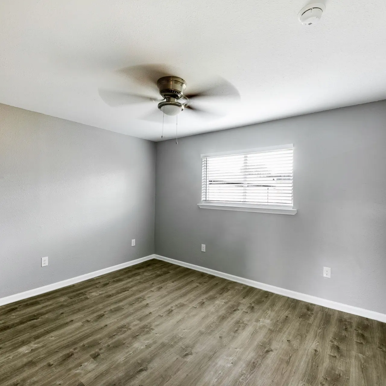 A vacant room featuring gray walls, a ceiling fan, and a window with blinds.