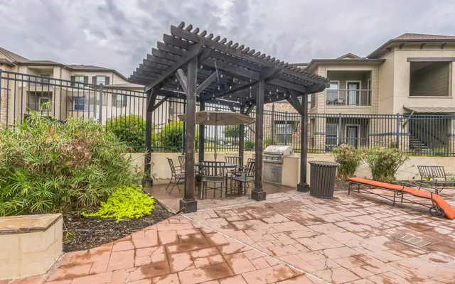 Outdoor Seating Area with Pergola Outdoor seating area with a pergola, table, and grill, surrounded by greenery and residential buildings.