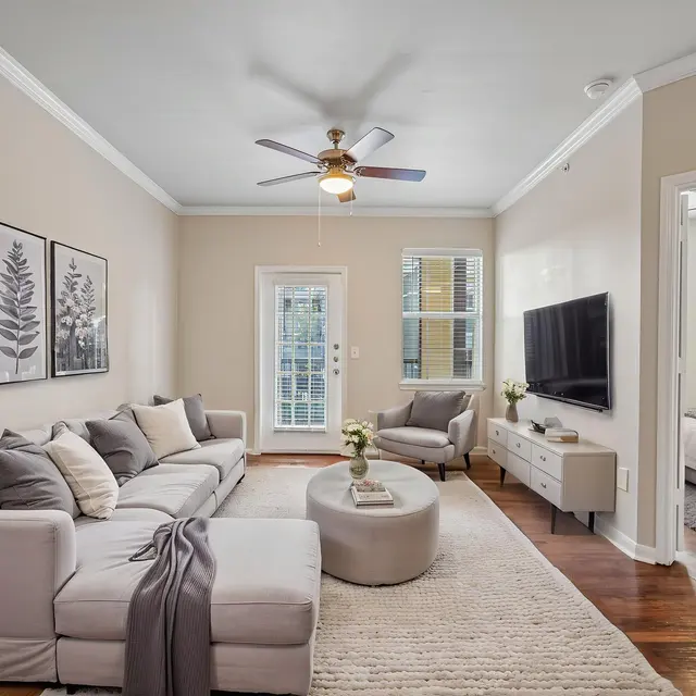 Living Room (Staged) - Tigoni Villas A modern living room featuring a gray sectional sofa with decorative pillows, a round coffee table, light walls, and framed artwork. A television is mounted on the opposite wall, and there are large windows allowing for natural light.
