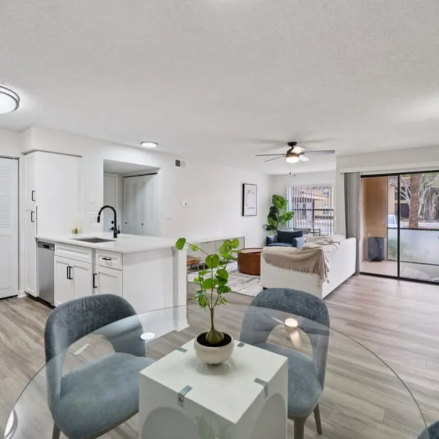 A beautifully designed modern apartment interior featuring an open layout with a dining area, kitchen, and living room. The dining table is set with four chairs, and the kitchen has white cabinets and a black refrigerator. A large sliding door leads to a patio area.