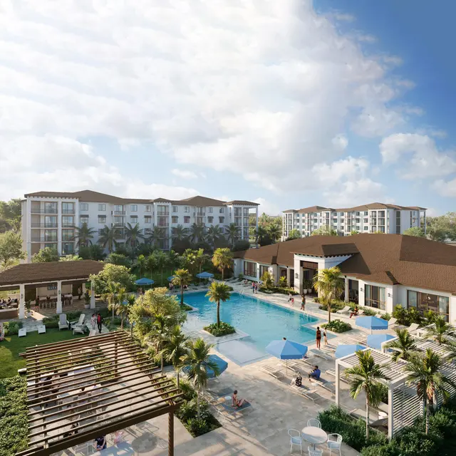 A modern resort featuring a large swimming pool surrounded by sun loungers, palm trees, and shaded areas. In the background, there are multi-story buildings and a lush green landscape.