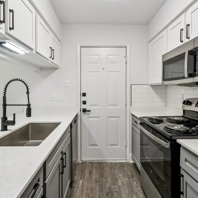 A modern kitchen featuring white cabinets, stainless steel appliances, and a large sink with a stylish faucet. The kitchen has a clean and bright look with an entrance door visible at the end.