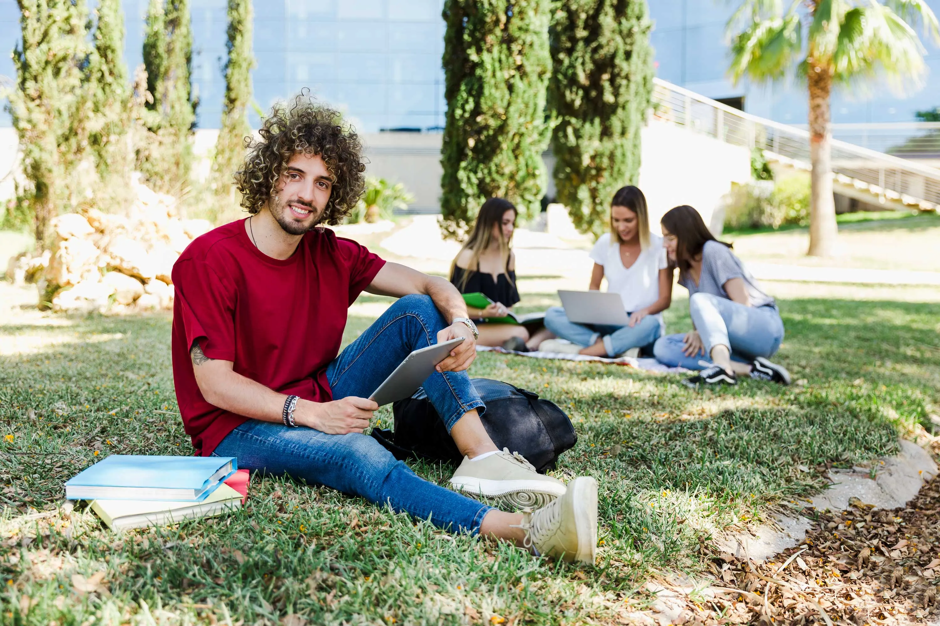Students Gathering in the Park A group of students sitting on the grass in a park. A young man in a red shirt is smiling at the camera while using a tablet. Three other female students are seen in the background, using laptops and sitting on a blanket.