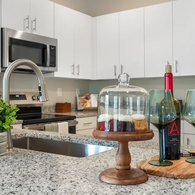 A modern kitchen featuring white cabinets, a stainless steel microwave, a granite countertop, and a sink. A glass dome with cupcakes is displayed on a wooden stand next to a bottle of wine and two green wine glasses on a wooden serving board. Greenery is placed in a pot on the counter.