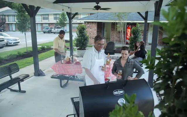 A backyard barbecue gathering with several people interacting. One man is grilling while a woman watches. A table with checkered cloth and drinks is nearby.