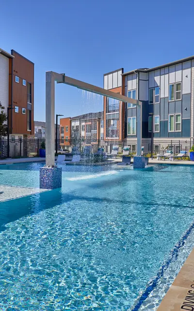 An outdoor swimming pool surrounded by modern apartment buildings. The pool features a waterfall structure and lounge chairs along the edge. The area is bathed in sunlight with clear blue skies above.