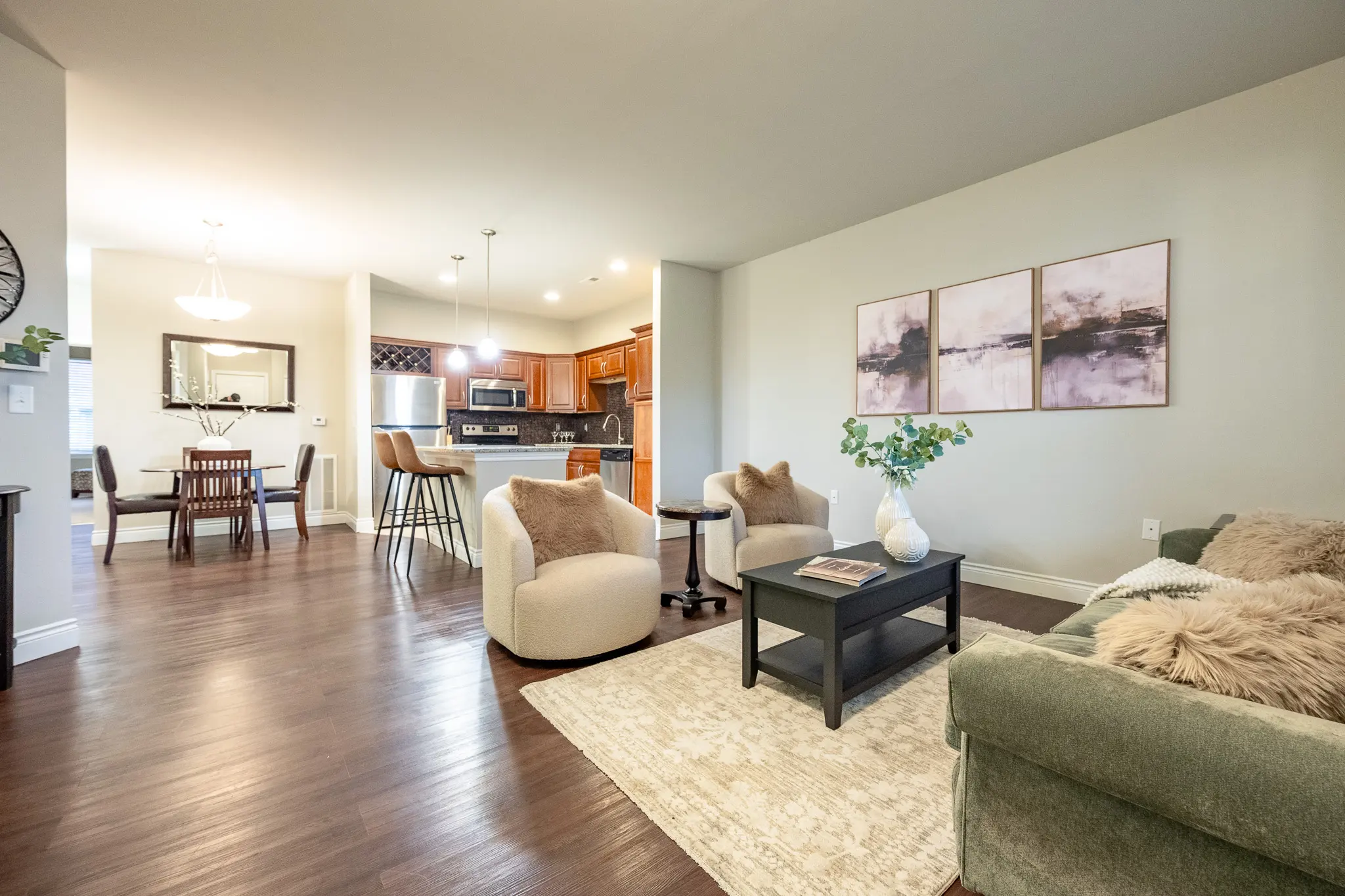 A stylish living room and dining area featuring modern furnishings and decor. Soft armchairs with throw pillows are positioned around a low coffee table on a patterned rug. The dining area includes a wooden table with chairs, and a glimpse of a kitchen can be seen in the background. The walls are painted in neutral tones, enhancing the brightness of the space.
