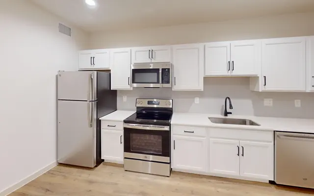 A modern kitchen featuring stainless steel appliances, including a refrigerator, microwave, oven, and dishwasher, with white cabinetry and a neutral-colored countertop. The floor is wooden and the space is brightly lit.