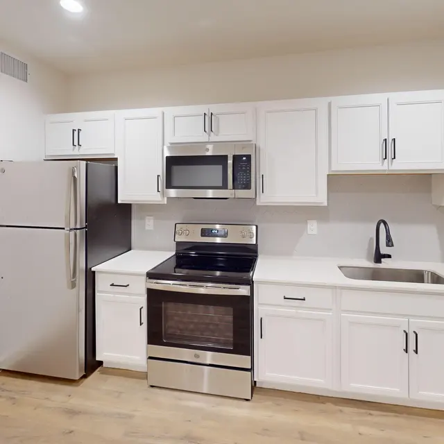 A modern kitchen featuring stainless steel appliances, including a refrigerator, microwave, oven, and dishwasher, with white cabinetry and a neutral-colored countertop. The floor is wooden and the space is brightly lit.