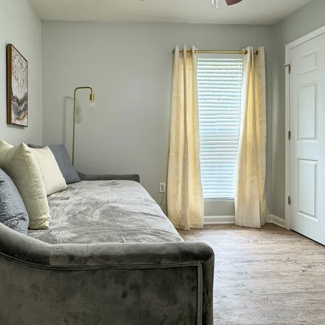 A cozy room featuring a gray sectional couch adorned with several decorative pillows, a standing lamp beside the couch, light-colored curtains hanging over a window, and framed artwork on the walls. The flooring is wooden.