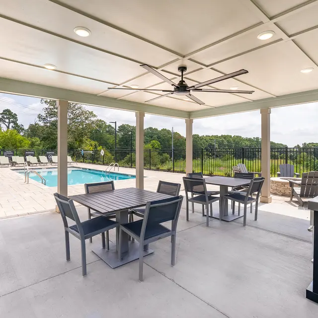 A covered outdoor dining area with a large table and chairs, next to a swimming pool surrounded by lounge chairs and green views.