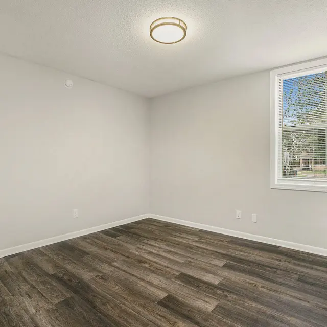A vacant room with light-colored walls, newly installed dark wood flooring, and a window allowing natural light. The ceiling has a modern light fixture, and there is no furniture present in the room.