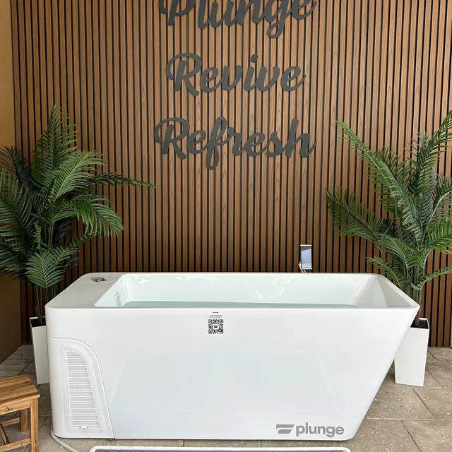 Spa Bathroom Setup with Bathtub A modern white bathtub with the word 'plunge' on it, set against a wooden wall with the words 'Plunge Revive Refresh' in large lettering above. There are potted plants on either side and a wooden stool in front.