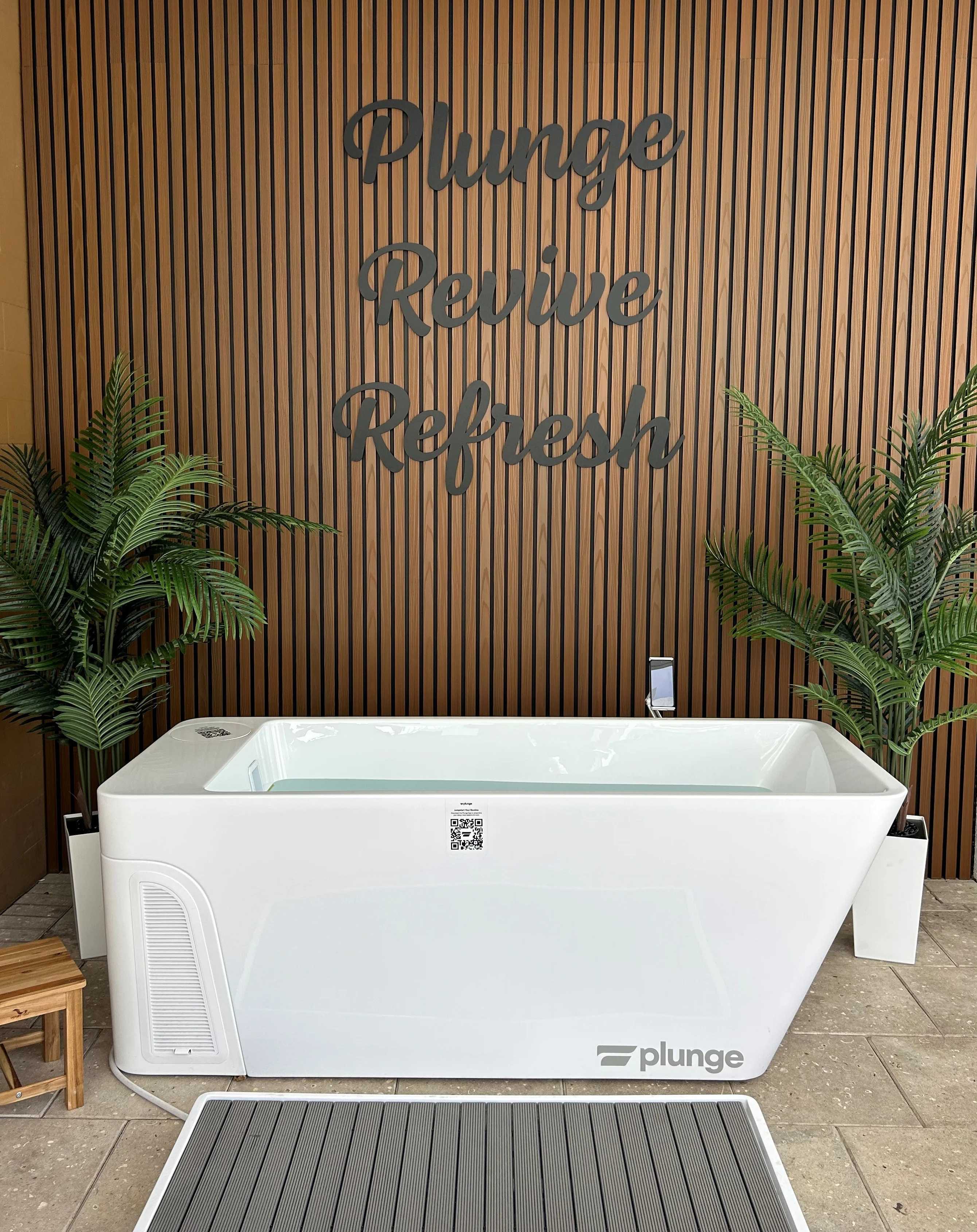 Spa Bathroom Setup with Bathtub A modern white bathtub with the word 'plunge' on it, set against a wooden wall with the words 'Plunge Revive Refresh' in large lettering above. There are potted plants on either side and a wooden stool in front.