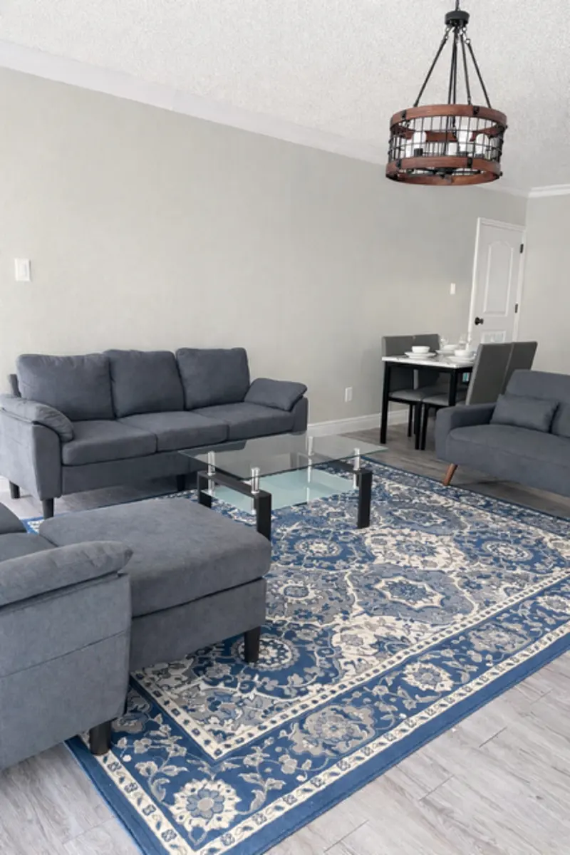 A modern living room featuring a gray sofa set, a glass coffee table, and a patterned blue rug. A dining area is visible in the background with a dining table and chairs. The decor is minimalist and contemporary.