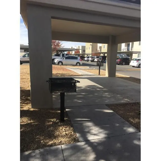 A barbecue grill in a small park area with a sidewalk leading toward a parking lot and apartments in the background.