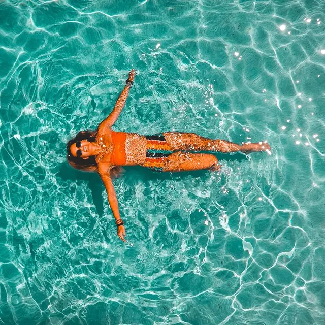 A woman floating on her back in a shimmering turquoise pool, surrounded by water reflecting light.