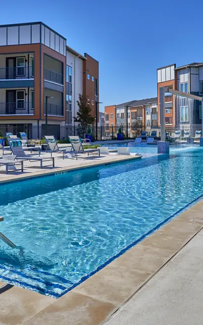 A modern apartment complex featuring a swimming pool surrounded by lounge chairs and trees, under a clear blue sky.