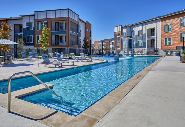 Apartment Pool Area A modern apartment complex featuring a swimming pool surrounded by lounge chairs and trees, under a clear blue sky.
