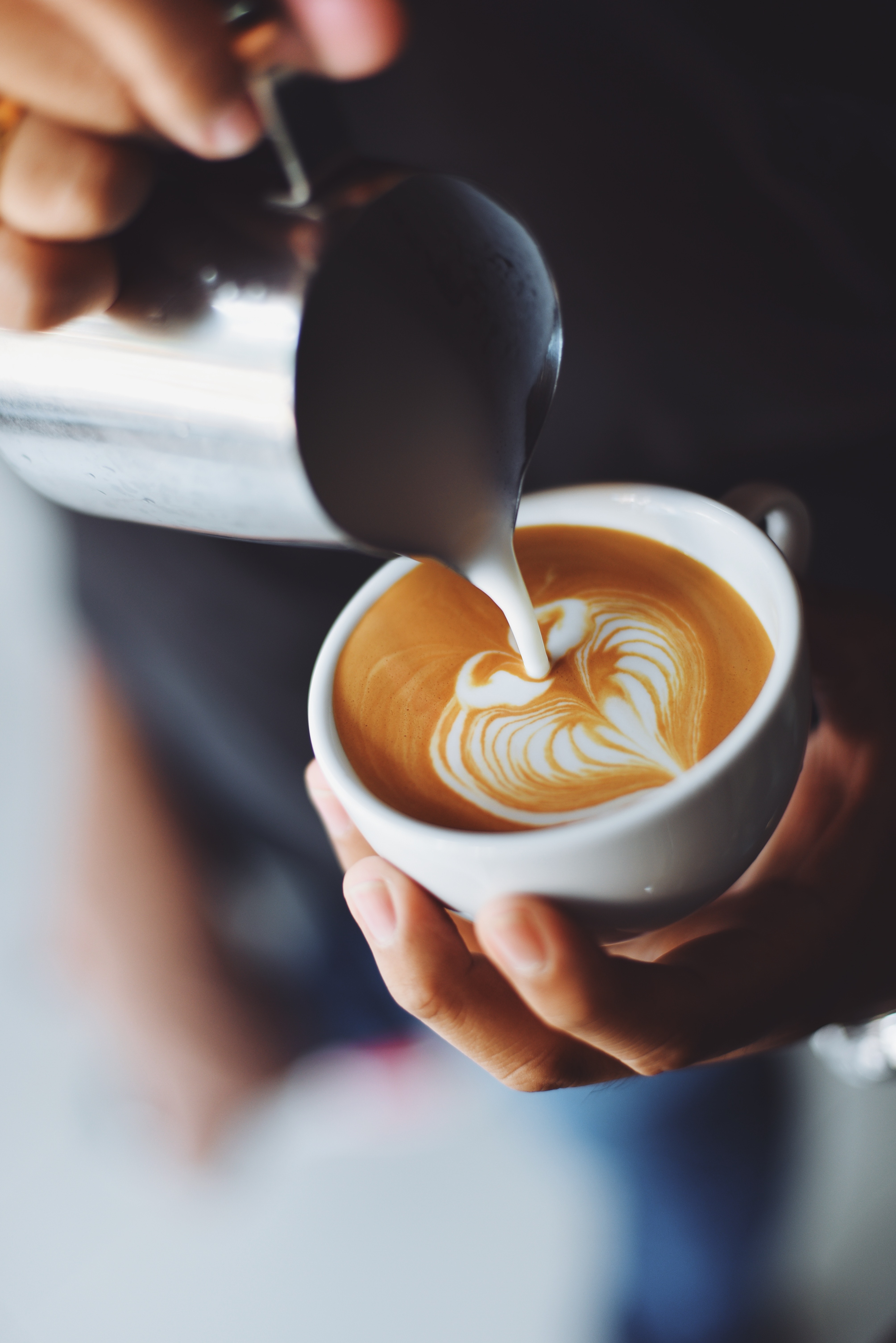 Latte Art in Progress A hand pouring steamed milk into a white cup of coffee, creating latte art in the shape of a heart. The background is softly blurred, focusing on the coffee cup.