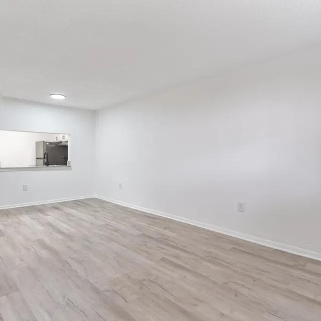 A spacious, empty living room with light-colored walls and wooden flooring. There is a doorway leading to another room on the left and an open kitchen area visible through an archway. The room is well-lit and has no furniture.