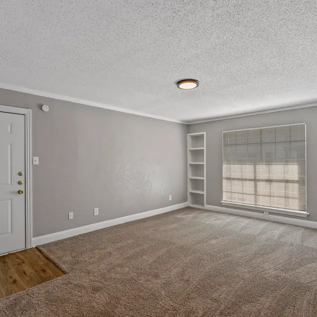 An empty living room featuring beige walls, carpet flooring, and a large window with blinds, along with a shelf on the wall and a front door.