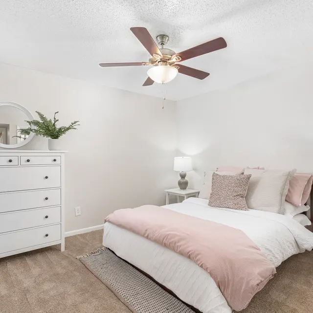 Spacious bedroom with carpeted floors, a ceiling fan, tall dresser beside the window, and two bedside tables.