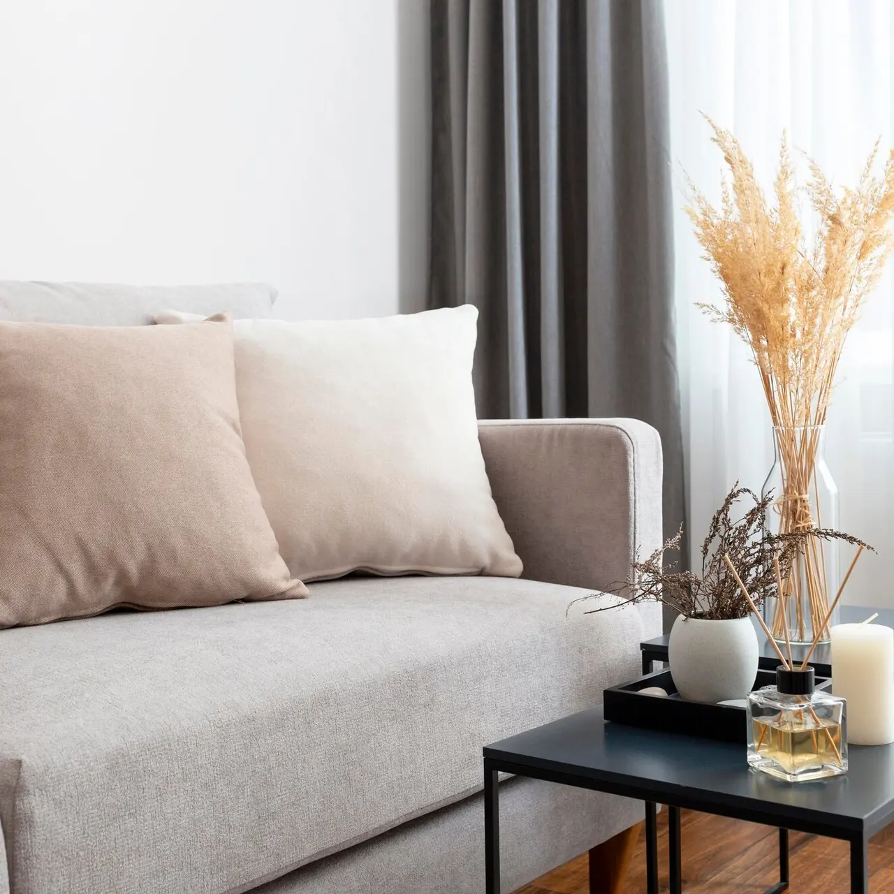 A gray sofa with beige pillows, next to a black table holding vases with dried flowers and a candle, against a window with gray curtains.