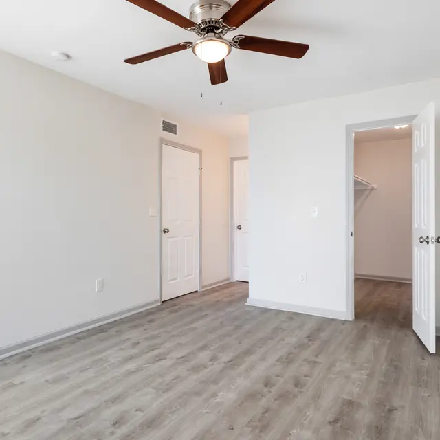 An empty room with light gray walls, a wooden floor, and a ceiling fan. One door is open, revealing another room. Two doors are visible on the left side of the room.