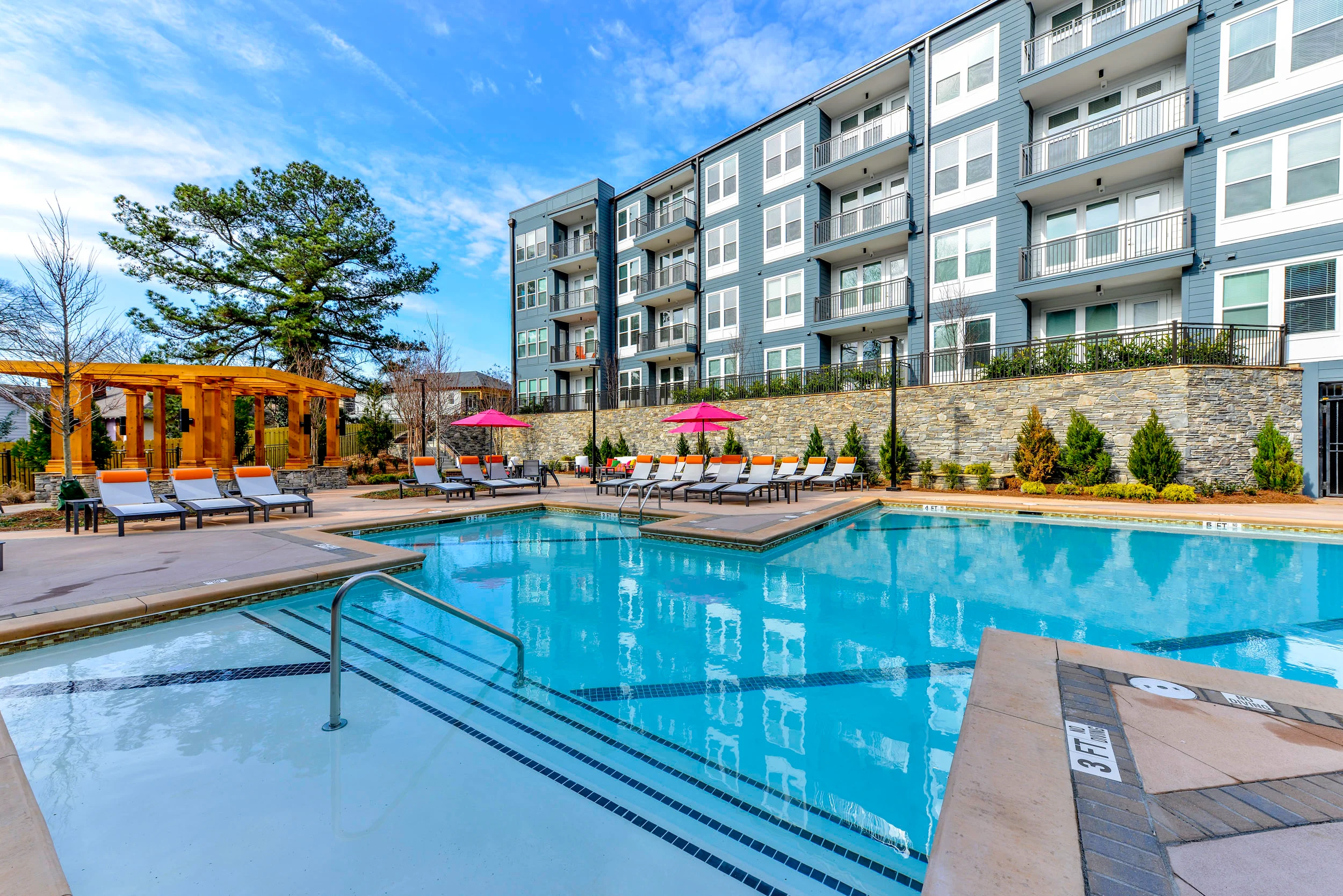 Pinnacle A modern apartment complex featuring a pool, lounge chairs, and umbrellas, surrounded by greenery and a clear blue sky.