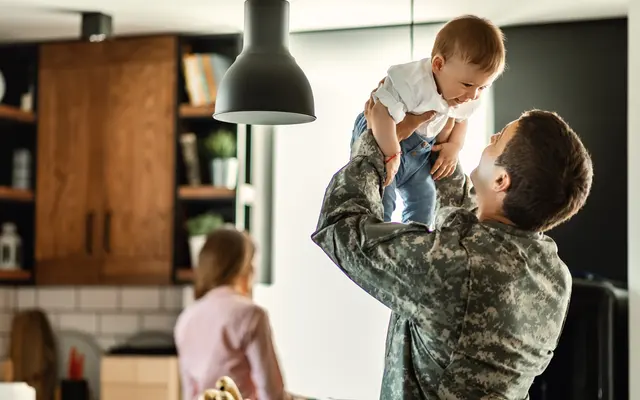 Oaks of Kingwood A person in military uniform lifts a smiling baby indoors, with another person standing in the kitchen background.