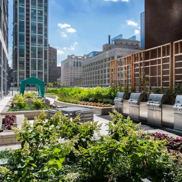 A modern urban rooftop garden featuring several stainless steel grills, surrounded by lush greenery and blooming flowers, with tall buildings in the background under a sunny sky.