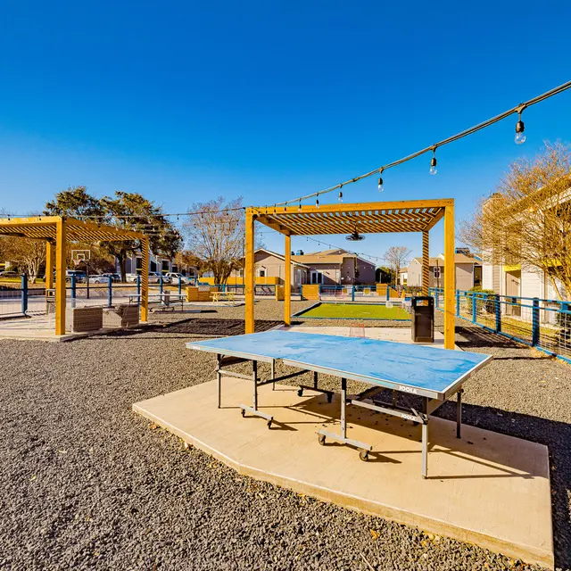 A spacious outdoor recreational area featuring a ping pong table on a concrete platform, surrounded by gravel and pergolas. The background shows basketball courts and nearby buildings under a clear blue sky.